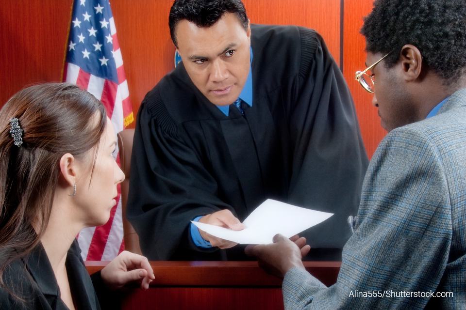 A judge accepts a paper from an attorney as another attorney looks on