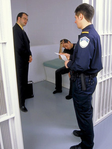 Photo of a jail cell, a lawyer is talking with a prisoner, with a police officer looking in. 