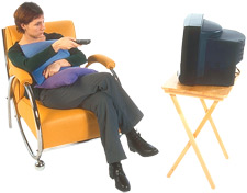 An early 2000's photo of a woman sitting in a chair, pointing a remote at a CRT TV on a wooden tableT