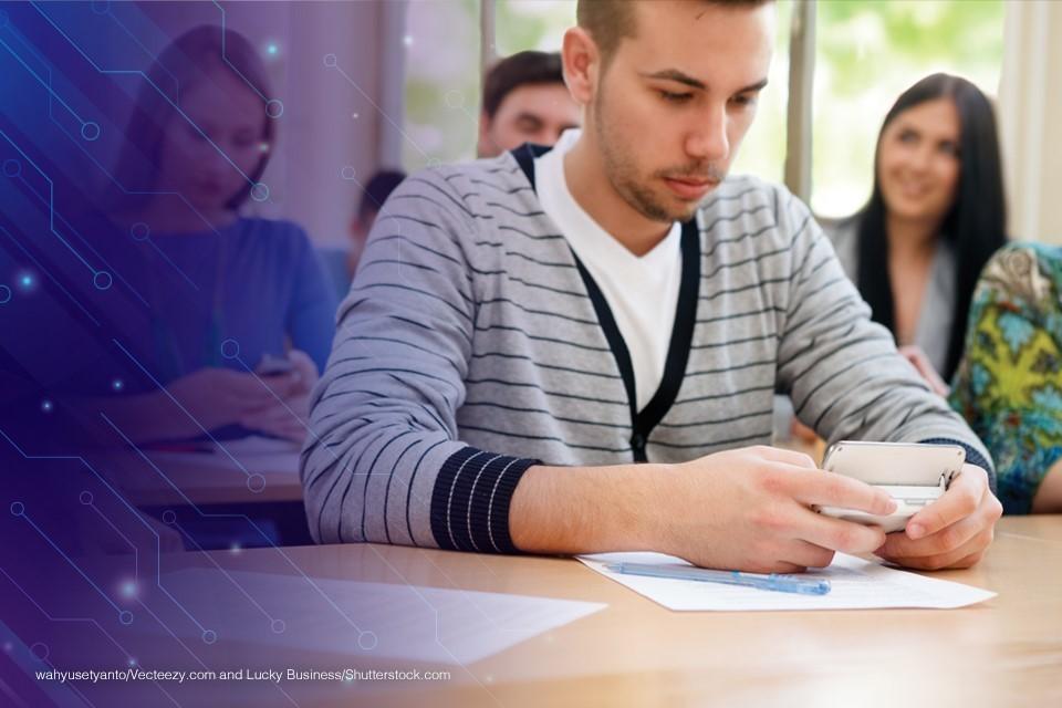 A classroom with a young person centered on their mobile device while classmates are talking with each other